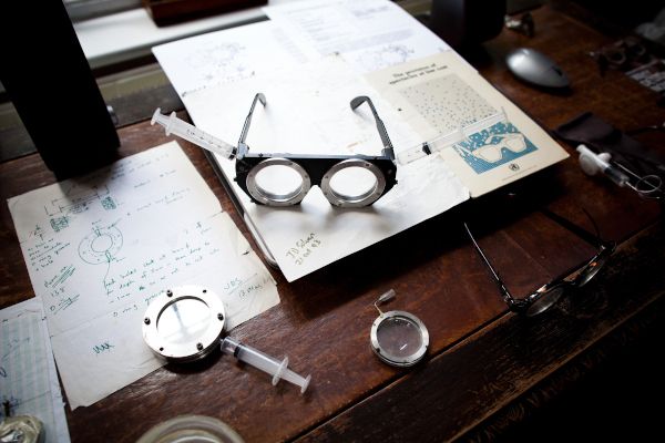 Eine selbstanpassende Brille liegt auf einem dunklen Schreibtisch auf Papier - zu sehen sind weitere Gegenstände