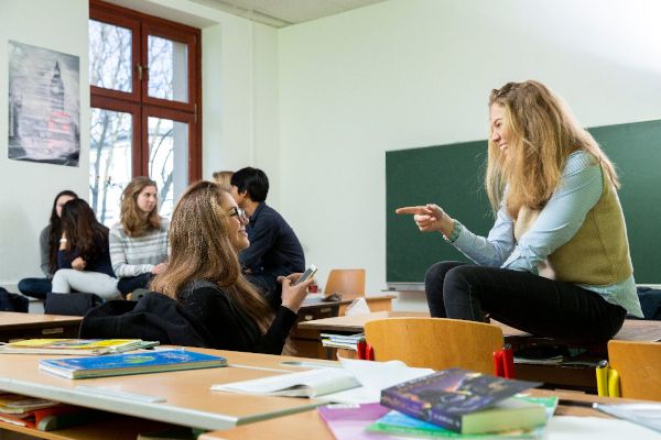 Zwei Schülerinnen sprechen miteinander in einem Klassenraum, im Hintergrund sind weitere Personen zu sehen