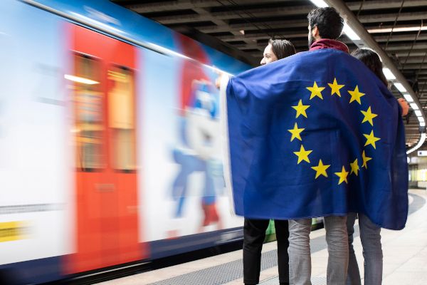 Drei Personen stehen im Kreis an einem Bahnsteig und sind von einer EU-Flagge umhüllt. Daneben ist ein vorbei fahrender Zug zu sehen.