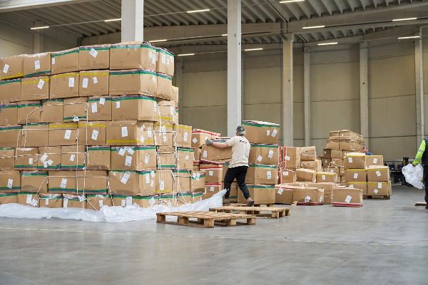 Ein Arbeiter in einer Lagerhalle voll mit Kartons, hebt ein Paket von einer Palette.
