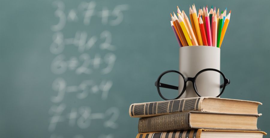 Foto: ein Stapel Bücher, darauf eine Dose mit Buntstiften und einer Brille. Im Hintergrund eine Schultafel mit Rechenbeispielen