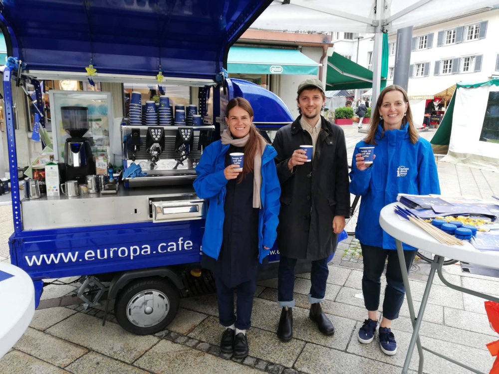 Drei Personen stehen vor der Europa Café-Ape und halten einen Kaffeebecher in der Hand