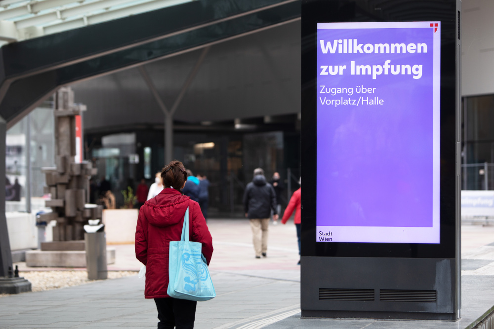 Menschen gehen neben einem Infoscreen für die Covid-19 Impfung vor dem Austria Center Wien