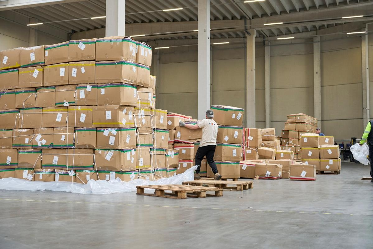Ein Arbeiter in einer Lagerhalle voll mit Kartons, hebt ein Paket von einer Palette.