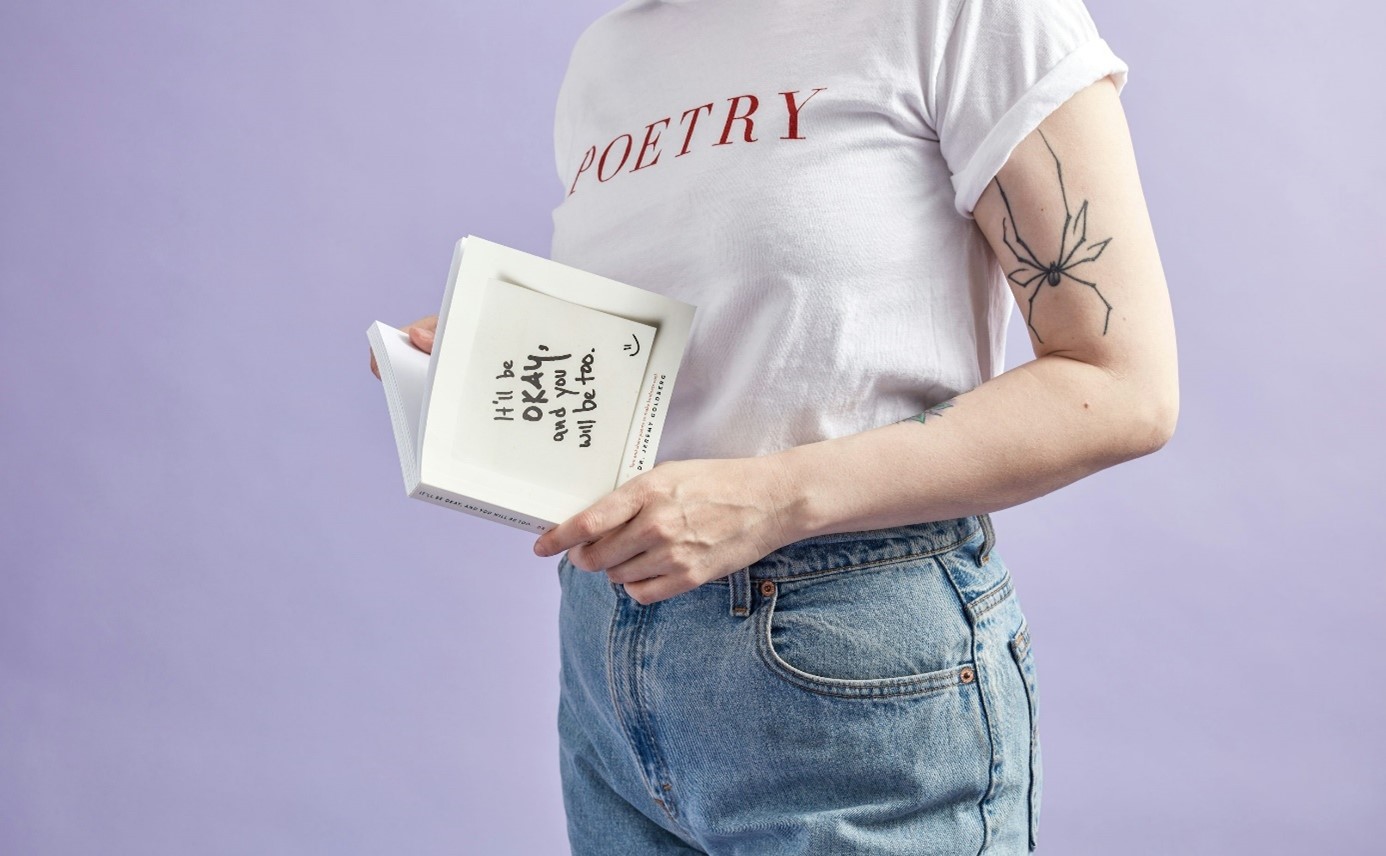 Frau mit T-Shirt mit Schriftzug "Poetry", Spinnentattoo am Oberarm, Notizblock in der Hand